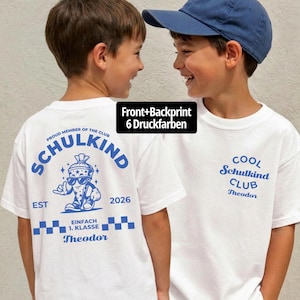 May include: A white t-shirt featuring blue graphic prints. The back displays a cartoon school cone character with sunglasses, the text "SCHULKIND," "EST 2026," and "EINFACH 1. KLASSE." The front features the text "COOL SCHULKIND CLUB" and "Theodor."