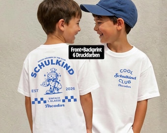 Puede incluir: Una camiseta blanca con estampados gráficos azules. La parte trasera muestra un personaje de cono escolar con gafas de sol, el texto "SCHULKIND", "EST 2026" y "EINFACH 1. KLASSE". La parte delantera tiene el texto "COOL SCHULKIND CLUB" y "Theodor".