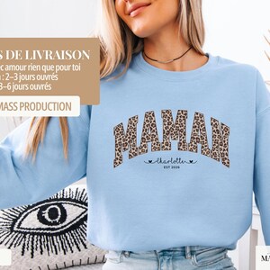 Op de afbeelding: Lichtblauw sweatshirt met het woord "MAMAN" in een luipaardprint. Onder het woord "MAMAN" staat de naam "Charlotte" en het jaar "EST 2026". Het sweatshirt is lichtblauw van kleur.