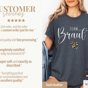 Pu&ograve; includere: T-shirt grigio scuro con la scritta "TEAM Braut" in corsivo bianco e un cuore con stampa leopardata. La maglietta ha un girocollo e maniche arrotolate. La maglietta fa parte della collezione Simply Marguerite.