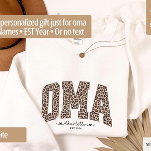 May include: White sweatshirt with the word "OMA" in a leopard print design. Below the word "OMA" is the name "Charlotte" and the year "EST 2026". The text "A personalized gift just for oma" is at the top.