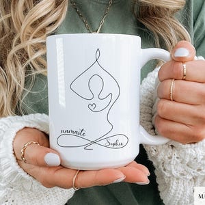 May include: White ceramic mug featuring a black line drawing of a yoga pose with a heart. The mug has the word "namaste" and the name "Sophie" in a script font. The mug is held by a person.
