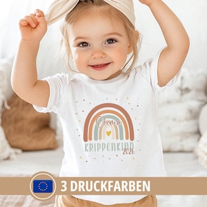 Op de afbeelding: Een wit T-shirt met korte mouwen en een afbeelding van een regenboog in gedempte tinten bruin, blauw en crème. De naam Olivia staat in cursief schrift in de regenboog, met de tekst KRIPPENKIND 2026 en een klein geel hartje eronder.
