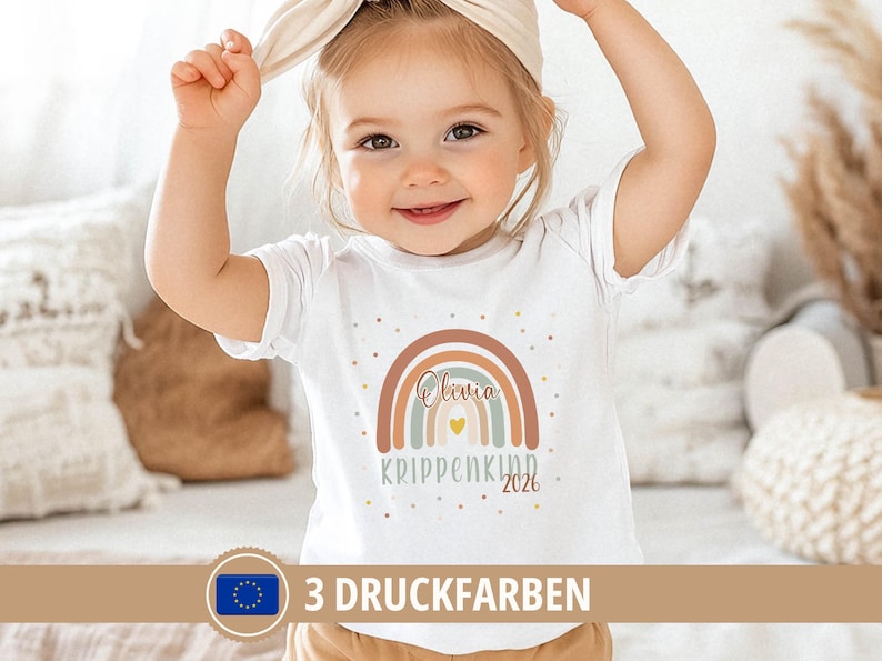 May include: A white short-sleeved t-shirt featuring a graphic of a multi-colored rainbow in muted tones of brown, blue, and cream. The name Olivia is printed in script inside the rainbow, with the text KRIPPENKIND 2026 and a small yellow heart below.