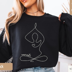 May include: Black sweatshirt featuring a white line drawing of a person in a yoga pose with the word "namaste" below. The design includes a heart shape. The words "super soft" are in the upper right corner.