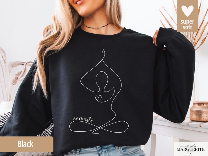 May include: Black sweatshirt featuring a white line drawing of a person in a yoga pose with the word "namaste" below. The design includes a heart shape. The words "super soft" are in the upper right corner.