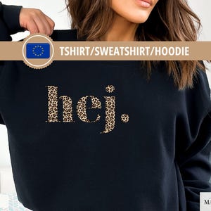May include: Black sweatshirt featuring the word "hej." in a leopard print pattern. The sweatshirt also has the text "TSHIRT/SWEATSHIRT/HOODIE" and the word "Black" on the bottom left. The brand name "simply MARGUERITE" is on the bottom right.