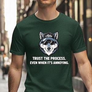 Peut inclure: T-shirt vert foncé avec un motif de loup portant des lunettes de soleil et une casquette avec le mot "Alpha". Le texte sur le t-shirt dit "Trust the process. Even when it's annoying." Le t-shirt est en matière souple.