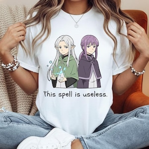 Può includere: T-shirt bianco con un'illustrazione in stile anime di due personaggi con capelli lunghi e mantelli. Il testo "This spell is useless." è stampato sotto l'immagine. La maglietta è indossata con jeans blu e scarpe da ginnastica bianche.