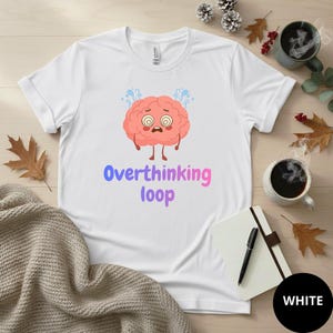 Può includere: T-shirt bianca con una grafica di un cervello dei cartoni animati e le parole "Overthinking loop" in blu e rosa. Il cervello ha un'espressione preoccupata. La maglietta è su una superficie di legno con tazze di caffè e un taccuino.