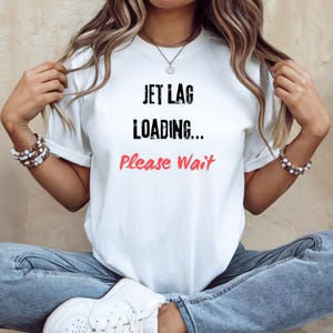 Può includere: T-shirt bianca con la scritta "JET LAG LOADING... Please Wait" in nero e rosso. La maglietta ha maniche corte ed è indossata da una persona. La persona indossa braccialetti e una collana.