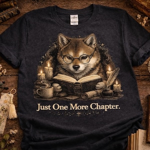 Può includere: T-shirt grigio scuro con un cane Shiba Inu con gli occhiali che legge un libro. Il design include candele, una penna e il testo "Just One More Chapter."