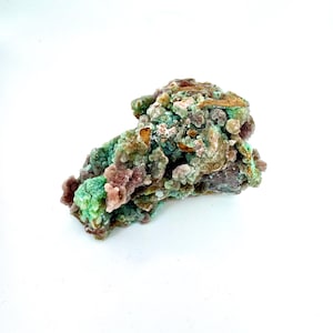 May include: A natural mineral specimen with a textured surface. The stone features a mix of colors, including green, brown, and purple. The mineral has a crystalline structure and is set against a white background.
