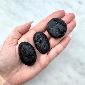 Könnte beinhalten: Drei kleine, schwarze, strukturierte Steine unterschiedlicher Form werden gezeigt. Einer ist rund, die anderen beiden oval. Die Steine haben eine raue, zerklüftete Oberfläche. Die Steine werden in einer offenen Hand vor einem weißen Marmorhintergrund gehalten.