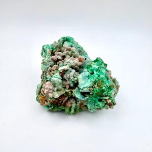 May include: A close-up of a natural mineral specimen. The rock features a vibrant green hue with areas of lighter green, brown, and pinkish-tan. The surface has a textured, crystalline appearance, with a rough, uneven shape.