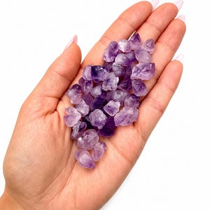 Pode incluir: Um punhado de pequenos cristais de ametista brutos em tons de roxo. Os cristais são segurados em uma mão aberta contra um fundo branco, mostrando suas formas naturais irregulares e sua qualidade translúcida. Estes cristais são populares para joias e usos metafísicos.