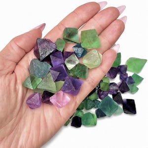 May include: A selection of fluorite crystals in shades of green, purple, and blue. The crystals are various sizes and shapes, with a mix of opaque and translucent surfaces. The crystals are scattered on a white background.