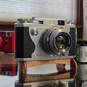 May include: A silver 35mm film camera with a black leather covering and a silver lens. The camera is sitting on a brown leather strap. The camera has the text "24x36" and "Hexanon 1:2 f=48mm" printed on it.