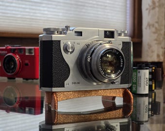 Puede incluir: Una cámara de película de 35 mm plateada con una cubierta de cuero negro y una lente plateada. La cámara está sentada sobre una correa de cuero marrón. La cámara tiene el texto "24x36" y "Hexanon 1:2 f=48mm" impreso en ella.