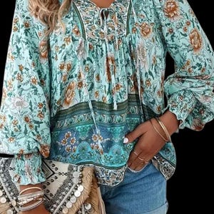 May include: A long-sleeved blouse in light turquoise featuring a multi-colored floral print, a V-neckline with tie strings, and smocked cuffs. The blouse is paired with blue denim shorts and a textured clutch bag adorned with metallic coin accents.