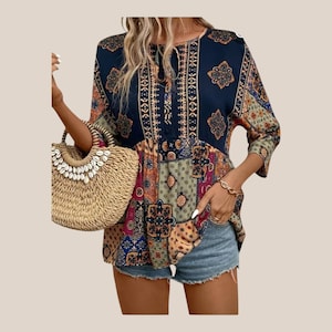 May include: A patchwork-style blouse with a round neckline and three-quarter sleeves. The top features a mix of patterns in navy, red, green, and gold. A straw tote bag with shell embellishments is held in the image. The model is wearing denim shorts.