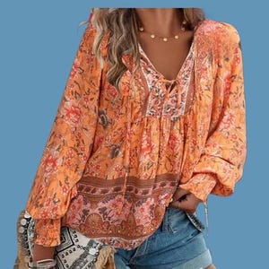 May include: Orange floral print blouse with long sleeves and a V-neck. The blouse features a tie detail at the neckline and a patterned border at the hem and cuffs. The fabric has a flowing, lightweight appearance, suitable for warm weather.