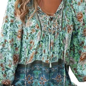 May include: A long-sleeved blouse with a floral pattern in shades of green, brown, and blue. The blouse features a V-neck with a tie closure and a contrasting blue floral border at the hem. The sleeves are long and loose.
