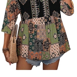 May include: A patchwork-style top with a black bodice and patterned sleeves. The top features a mix of green, orange, and beige geometric designs. The neckline has a tie detail. The model is wearing denim shorts and carrying a straw bag.