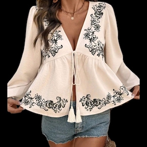 May include: Cream-colored blouse with long sleeves and a deep V-neck. The blouse features black floral embroidery along the neckline, hem, and sleeves. It has a tie closure with tassels and a peplum-style silhouette. The blouse is paired with denim shorts.