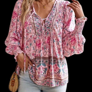 May include: Pink floral print blouse with a V-neckline and long sleeves. The blouse features a tie detail at the neckline and elastic cuffs. The floral pattern includes pink, green, and brown accents. The sleeves are flared at the wrist.