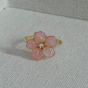 Puede incluir: Un anillo dorado con un diseño de flor rosa y un centro de piedra preciosa transparente. La flor tiene cinco pétalos con un acabado brillante. El anillo está sobre un fondo neutro.