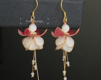 Puede incluir: Un par de pendientes florales hechos a mano. Cada pendiente presenta un diseño blanco en forma de pétalo con un acento rojo, alambre dorado y cuentas colgantes. Los pendientes están suspendidos de ganchos dorados, creando un aspecto delicado.