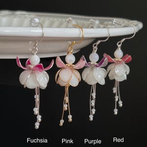 Può includere: Quattro orecchini a fiori fatti a mano fucsia, rosa, viola e rossi. Ogni orecchino presenta un design a fiore bianco con un filo di perline pendenti. Gli orecchini sono esposti su una superficie bianca. Il testo sull'immagine recita "Fuchsia", "Pink", "Purple" e "Red".