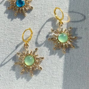 Puede incluir: Pendientes dorados con forma de sol con una gema verde central. Los pendientes tienen cierre de palanca y se exhiben sobre una superficie texturizada de color gris claro. Yukidesign.