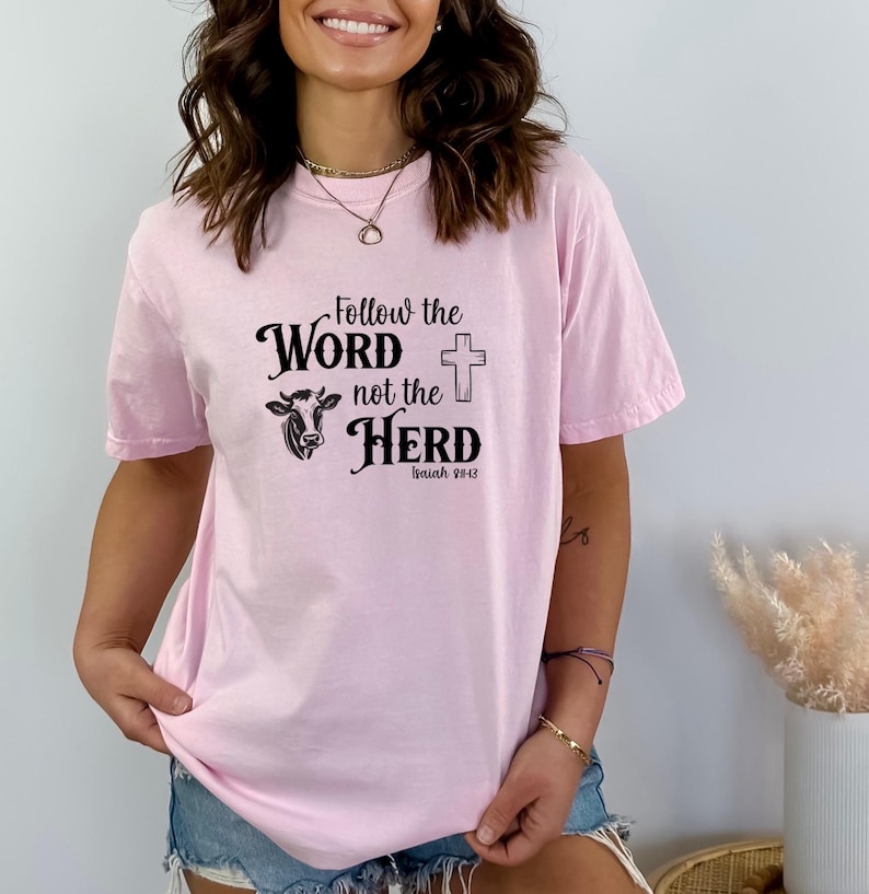May include: A light pink t-shirt with the text "Follow the Word not the Herd" in black script, a cow graphic, and a cross. The text "Isaiah 28:13" is below the main text. The shirt is a casual style.