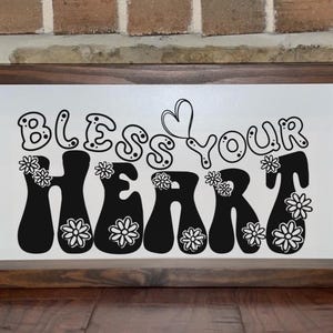 May include: A framed wooden sign with the phrase "Bless Your Heart" in black, groovy font. The word "Heart" is decorated with white daisy-like flowers. The background is white, and the frame is dark brown.