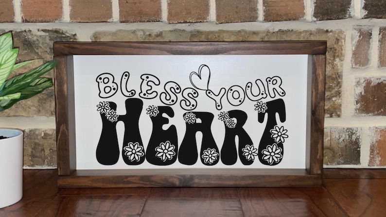 May include: A framed wooden sign with the phrase "Bless Your Heart" in black, groovy font. The word "Heart" is decorated with white daisy-like flowers. The background is white, and the frame is dark brown.