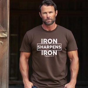 May include: A brown t-shirt with the text "IRON SHARPENS IRON" in a white, blocky font. The text is centered and framed by a white rectangle. Below the text is the scripture reference "Proverbs 27:17".