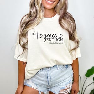 May include: Cream-colored t-shirt with the text "His grace is enough" in a black script font. Below the text is "2 Corinthians 12:9". The shirt is paired with light blue denim shorts.