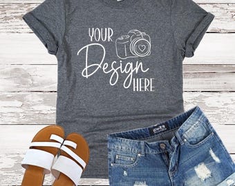 May include: A dark gray t-shirt with rolled sleeves, denim shorts, and sandals. The t-shirt features the text "YOUR Design HERE" with a camera graphic. The sandals have brown soles and white straps.
