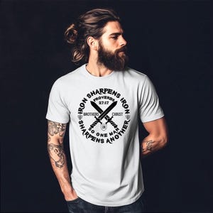 May include: A pale grey t-shirt with a black graphic. The design includes two crossed swords, the text "Iron Sharpens Iron" in a circular pattern, and the Bible verse Proverbs 27:17. The shirt has short sleeves.