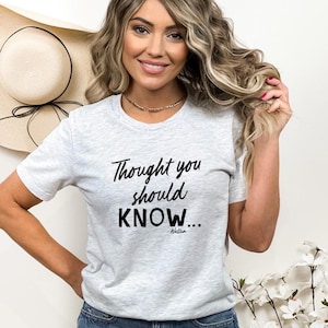 May include: A light gray t-shirt with the text "Thought you should KNOW..." in black script. The shirt is short-sleeved and has a crew neck. A straw hat with a black ribbon is visible in the background.