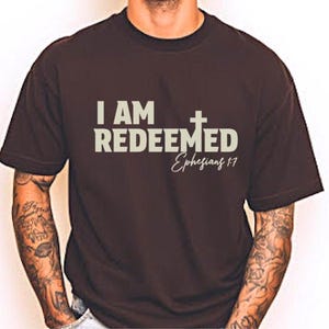 May include: A dark brown t-shirt with the text "I AM REDEEMED" in a light grey font, accompanied by a cross symbol and the inscription "Ephesians 1:7". The shirt is short-sleeved.