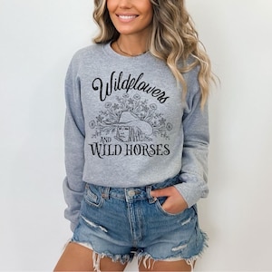 May include: A light gray sweatshirt with the text "Wildflowers and Wild Horses" in black script and a graphic of a woman wearing a hat. The model is wearing denim shorts.