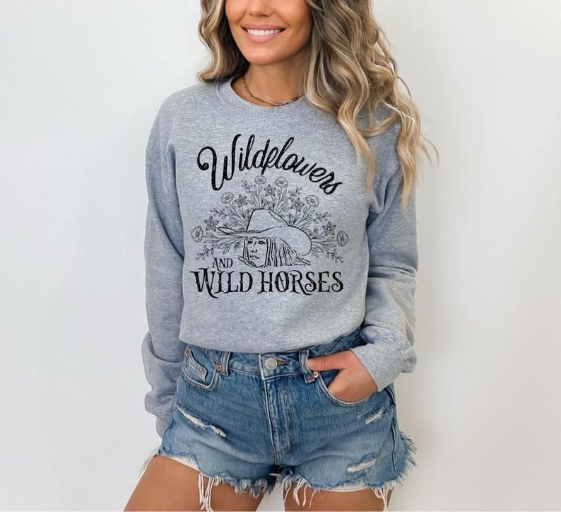 May include: A light gray sweatshirt with the text "Wildflowers and Wild Horses" in black script and a graphic of a woman wearing a hat. The model is wearing denim shorts.