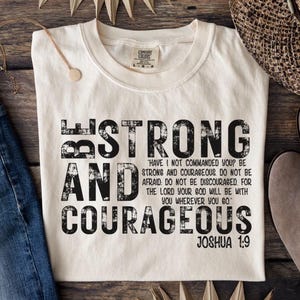 Könnte beinhalten: Cremefarbenes T-Shirt mit den Worten "BE STRONG AND COURAGEOUS" in großen, schwarzen, verwaschenen Buchstaben. Der Text enthält einen Bibelvers aus Josua 1:9. Das Shirt wird mit einer Halskette, Jeans und Stiefeln präsentiert.