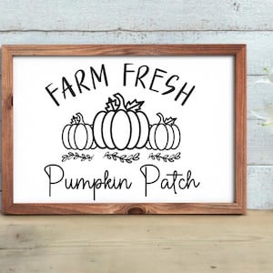 May include: A framed sign with the words "FARM FRESH Pumpkin Patch" in black script. The design includes three pumpkin illustrations and leafy accents. The frame is made of wood and is displayed on a wooden surface.