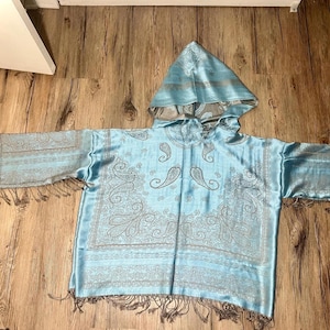 May include: A light blue hooded poncho with a paisley pattern in silver. The poncho has long sleeves with fringe at the ends and a matching hood. The fabric has a silky sheen.