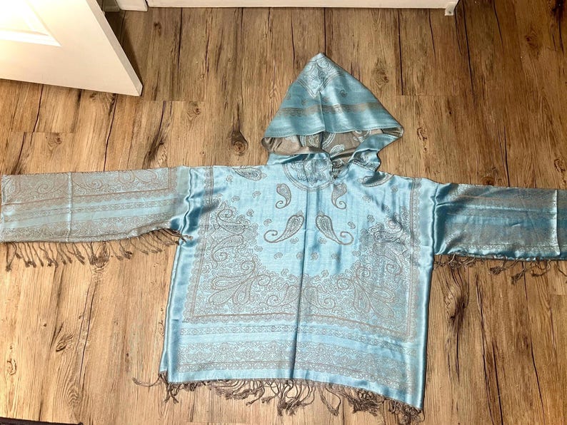 May include: A light blue hooded poncho with a paisley pattern in silver. The poncho has long sleeves with fringe at the ends and a matching hood. The fabric has a silky sheen.