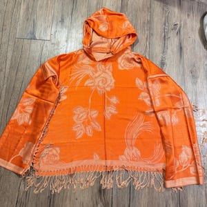 May include: An orange hooded scarf with a floral rose pattern. The scarf has a fringe along the bottom edge and is made of a soft, flowing material. The hood is attached to the scarf, creating a cozy and stylish accessory.